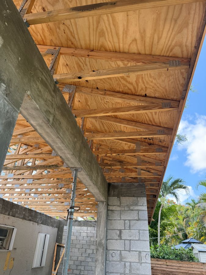 Residential addition in progress in Miami-Dade: concrete beam bearing on a concrete block column, with wood roof trusses and plywood roof sheathing above and a temporary steel shore post