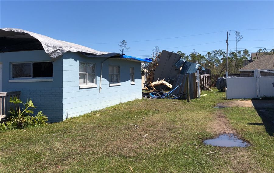 Single-story concrete block home in Florida with a damaged roof covered by tarps and debris piled in the side yard